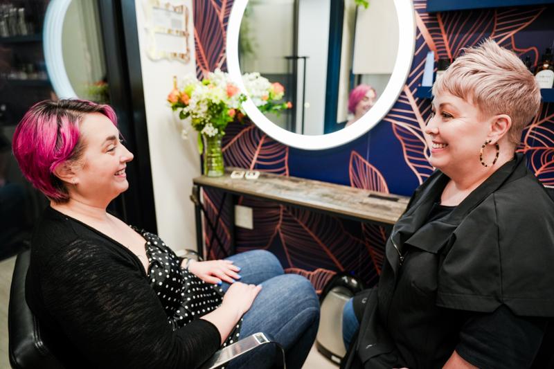 Hairstylist and client with bright pink hair sitting down in a consultation in the salon