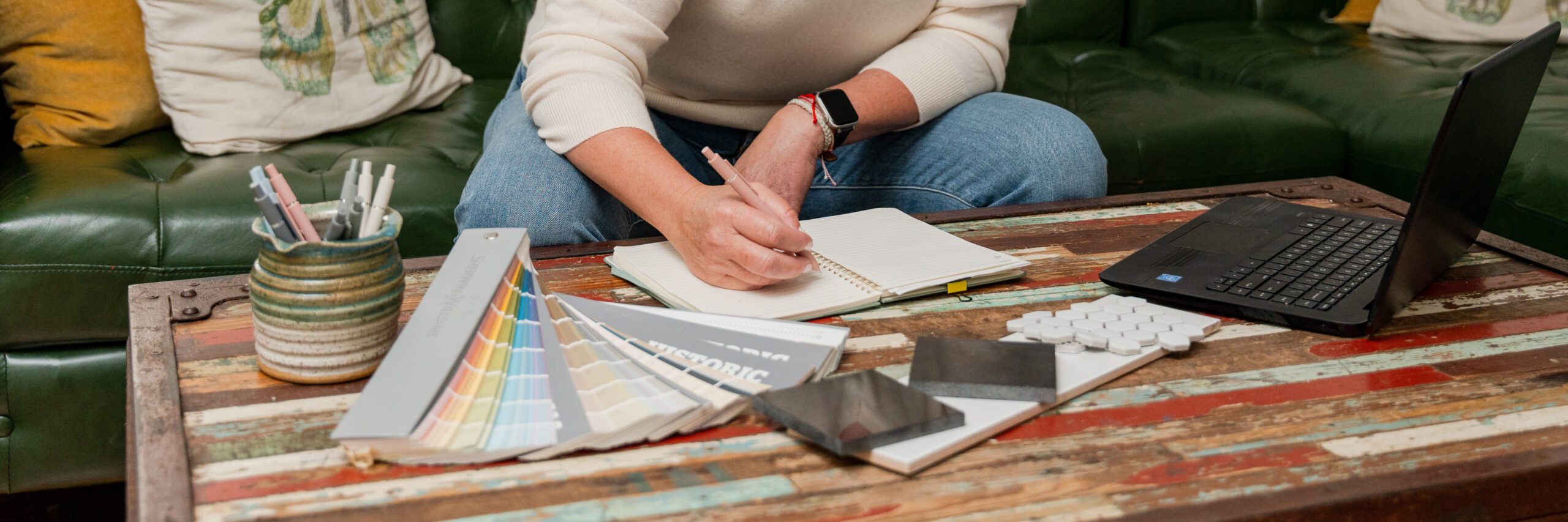 Designer with paint and finishes spread out on the table is making notes in their workbook.