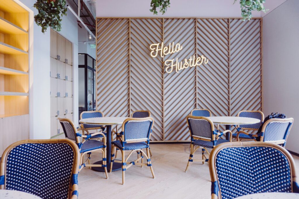 Coworking space with round tables and blue chairs. Accent wall with a neon sign says "Hello Hustler".