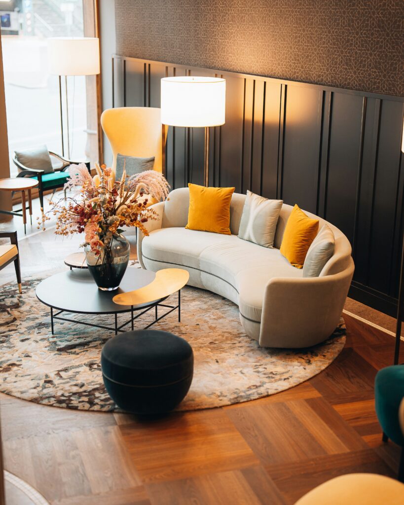 Modern and moody hotel lobby with a beige rounded couch, yellow pillows a round coffee table, round floor rug against a dark wall.