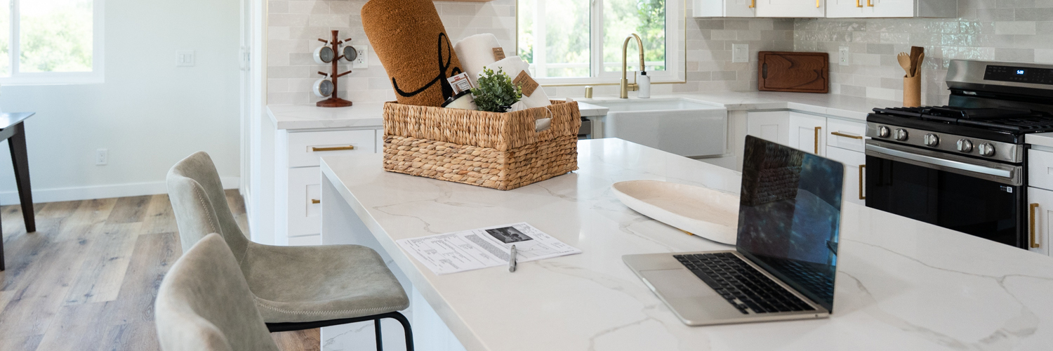 White kitchen countertop with bar stools. Laptop is open and some real estate documents are on the counter with a gift basket.
