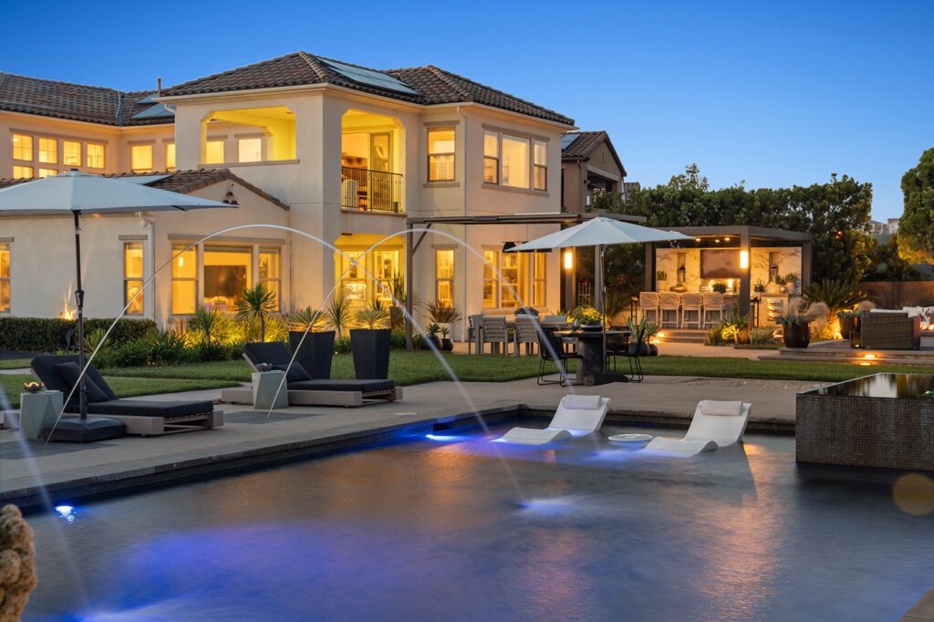 Twilight of a large two story home brightly lite overlooking a pool and well landscaped backyard aera.